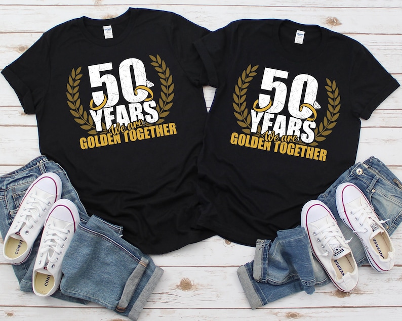 May include: Two black t-shirts with gold lettering and a gold laurel wreath design. The text on the shirts reads "50 Years We are Golden Together".