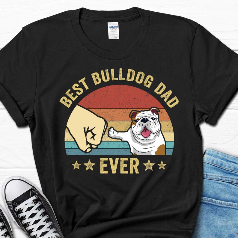May include: Black t-shirt with a vintage-style graphic that says "Best Bulldog Dad Ever" with a bulldog paw bumping a fist. The graphic has a sunset background with red, orange, and yellow stripes.