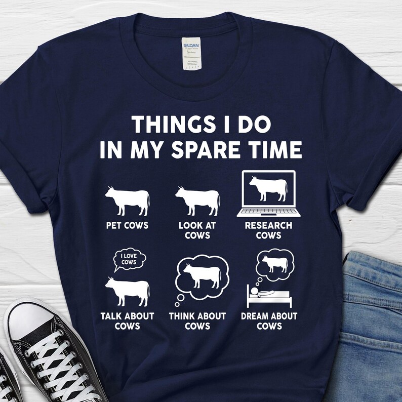 以下が含まれることがあります： 「Things I do in my spare time」という白い文字が書かれた紺色のTシャツ。テキストの後に、牛のイラストが6つあり、それぞれ「Pet Cows」、「Look at Cows」、「Research Cows」、「Talk about Cows」、「Think about Cows」、「Dream about Cows」というテキストが添えられています。