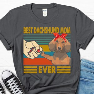 May include: A dark gray t-shirt with a vintage-style graphic of a dachshund wearing a red bandana. The text "Best Dachshund Mom Ever" is printed on the shirt.