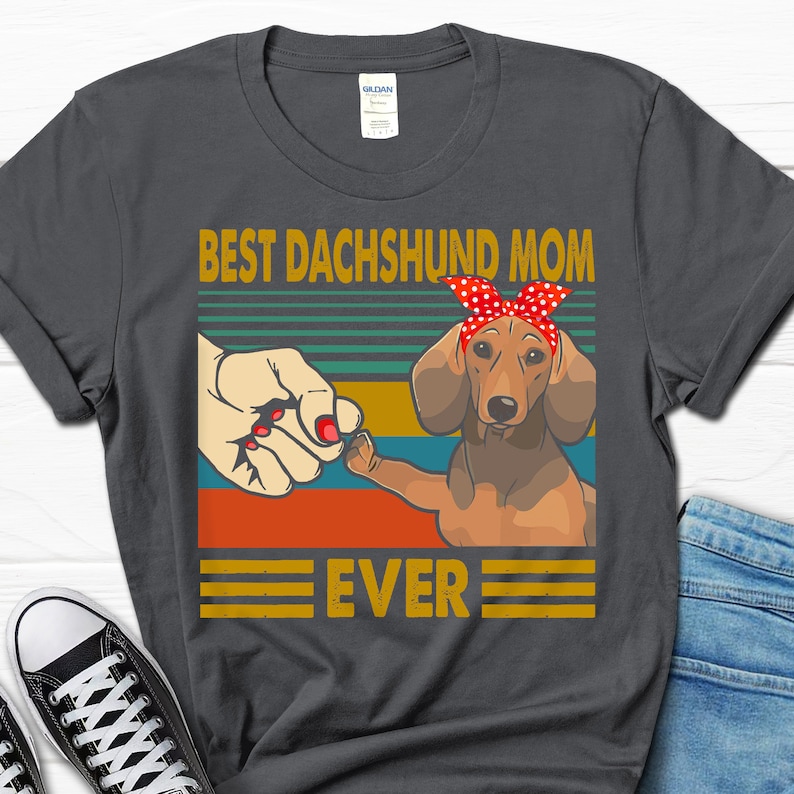 May include: A dark gray t-shirt with a vintage-style graphic of a dachshund wearing a red bandana. The text "Best Dachshund Mom Ever" is printed on the shirt.