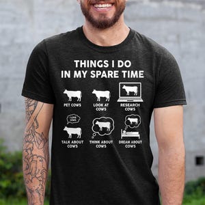 以下が含まれることがあります： 「Things I do in my spare time」と書いてある白いグラフィックが施された黒いTシャツ。牛が様々な活動をしている様子が描かれており、牛をなでたり、見たり、調べたり、話したり、考えたり、夢を見たりしている様子が描かれている。