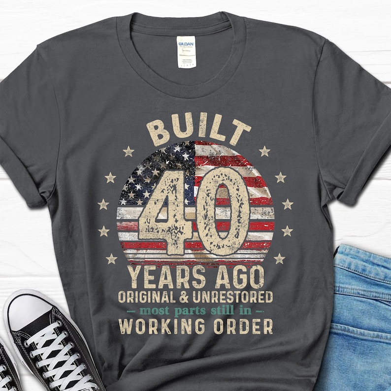 May include: A dark gray t-shirt with a vintage-style graphic design. The graphic features a red, white, and blue American flag with the number 40 in the center. The text "Built 40 Years Ago Original & Unrestored - most parts still in - Working Order" is printed below the flag.
