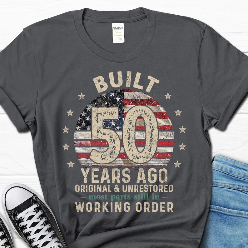 May include: A dark grey t-shirt with a vintage-style graphic design. The graphic features a red, white and blue American flag with the number 50 in the centre. The text "BUILT 50 YEARS AGO ORIGINAL & UNRESTORED - most parts still in - WORKING ORDER" is printed below the flag.