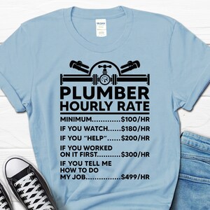 May include: Light blue t-shirt with a black graphic of a plumber's tools and the text "Plumber Hourly Rate". The graphic includes a wrench, pipe, and valve. The text below the graphic lists different hourly rates for a plumber based on the level of assistance provided.  The rates are: Minimum $100/hr, If you watch $180/hr, If you "help" $200/hr, If you worked on it first $300/hr, If you tell me how to do my job $499/hr.