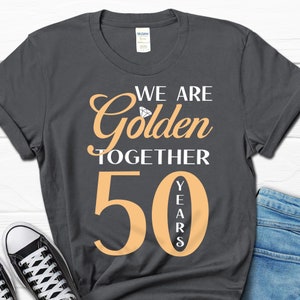 May include: A dark gray t-shirt with the text "We are Golden Together 50 Years" in white and orange. The number 50 is large and orange, with the word "Years" vertically stacked inside the number.