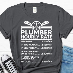 May include: A dark gray t-shirt with a white graphic of a plumber's tools and the text "Plumber Hourly Rate". The graphic includes a pipe with a valve and two wrenches. The text below the graphic lists different hourly rates for a plumber based on the level of assistance provided. The rates are: $100/hr for minimum, $180/hr for watching, $200/hr for helping, $300/hr for working on it first, and $499/hr for telling the plumber how to do their job.