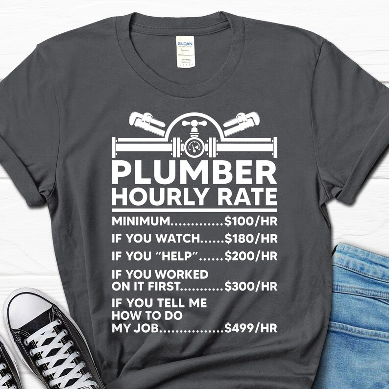 May include: A dark gray t-shirt with a white graphic of a plumber's tools and the text "Plumber Hourly Rate". The graphic includes a pipe with a valve and two wrenches. The text below the graphic lists different hourly rates for a plumber based on the level of assistance provided. The rates are: $100/hr for minimum, $180/hr for watching, $200/hr for helping, $300/hr for working on it first, and $499/hr for telling the plumber how to do their job.