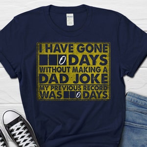 Op de afbeelding: Een marineblauw T-shirt met een gele afbeelding waarop staat: "Ik ben 0 dagen zonder een vaders grapje gegaan. Mijn vorige record was 0 dagen."