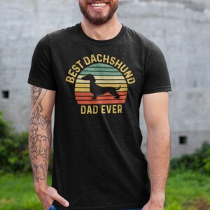 May include: A black t-shirt with a vintage-style graphic of a dachshund silhouette in front of a sunset. The text "Best Dachshund Dad Ever" is printed below the graphic.