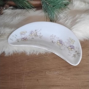 Puede incluir: Un plato blanco en forma de media luna con un estampado floral en tonos morado, amarillo y verde. El plato tiene una forma ligeramente curvada y un diseño delicado, perfecto para servir aperitivos o porciones pequeñas.