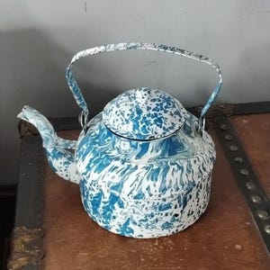 Op de afbeelding: Een blauwe en witte emaille theepot met een marmerpatroon. De theepot heeft een rond lichaam en een tuit aan de zijkant. De handgreep is bevestigd aan de bovenkant van de theepot.