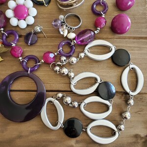 Op de afbeelding: Een paarse en zilveren ketting met grote ovale en ronde kralen. De ketting heeft een grote paarse hanger met een cirkel uitgesneden uit het midden. De ketting heeft ook zwarte en zilveren kralen.