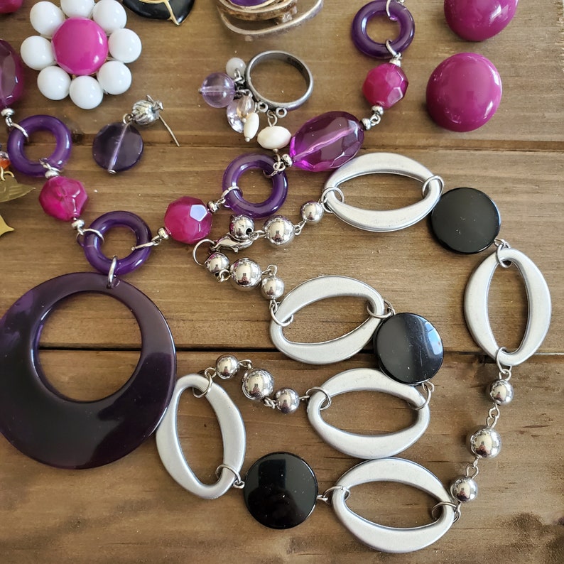 Op de afbeelding: Een paarse en zilveren ketting met grote ovale en ronde kralen. De ketting heeft een grote paarse hanger met een cirkel uitgesneden uit het midden. De ketting heeft ook zwarte en zilveren kralen.