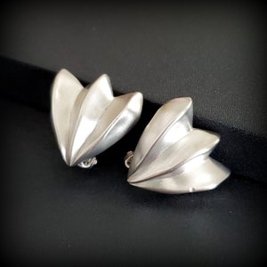 May include: A pair of silver-toned clip-on earrings. Each earring features a layered, petal-like design with a smooth, reflective surface. The earrings are set against a dark background, highlighting their metallic sheen and sculptural form.