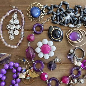 Op de afbeelding: Een verzameling van paarse, roze en witte kralen, samen met gouden en zilveren bedels, zijn gerangschikt op een houten oppervlak. De kralen zijn geregen aan kettingen en armbanden, en de bedels omvatten een bloem, een ster en een hart.