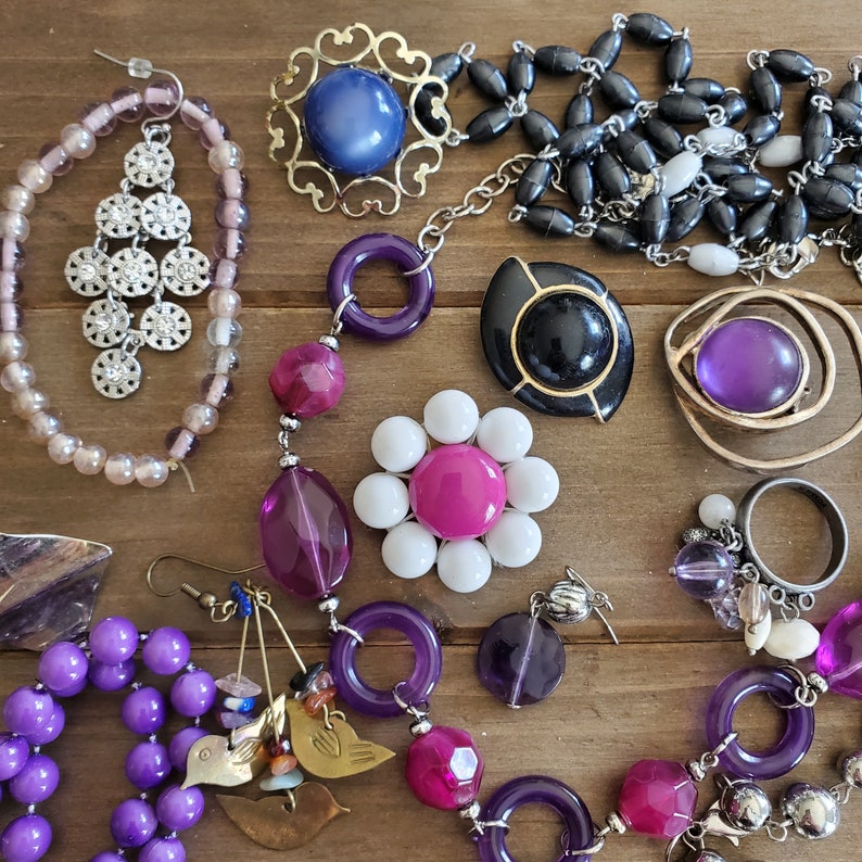 Op de afbeelding: Een verzameling van paarse, roze en witte kralen, samen met gouden en zilveren bedels, zijn gerangschikt op een houten oppervlak. De kralen zijn geregen aan kettingen en armbanden, en de bedels omvatten een bloem, een ster en een hart.