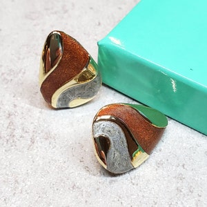 May include: A pair of abstract earrings with a gold trim. The earrings feature a teardrop shape with sections of shimmering copper, gray, and green enamel. The earrings are set against a light gray surface, with a turquoise box in the background.