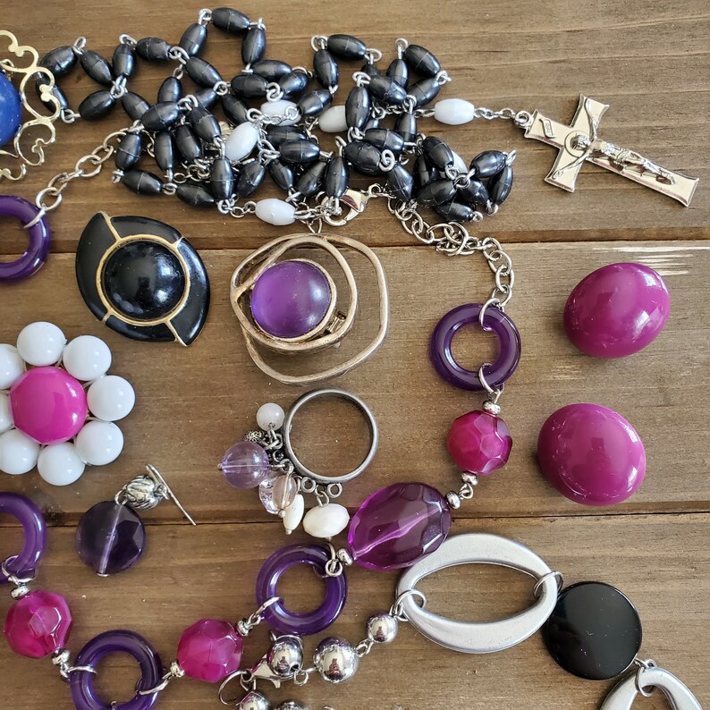 Op de afbeelding: Een verzameling sieraden met paarse, zwarte en witte kralen, bedels en hangers. De sieraden omvatten een ketting met een kruis hanger, een armband met verschillende kralen en een ring met een heldere steen.
