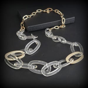 May include: A necklace featuring a combination of clear acrylic and gold-tone metal chain links. The necklace is displayed on a black surface, showcasing its design and materials. The chain links are oval-shaped.
