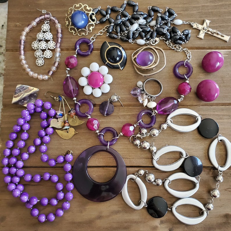 Op de afbeelding: Een verzameling sieraden met paarse, witte en zwarte kralen, een zilveren ketting met een kruisbedel en verschillende bedels, waaronder bloemen, sterren en geometrische vormen.