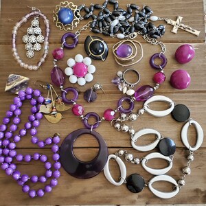 Op de afbeelding: Een verzameling sieraden met paarse, witte en zwarte kralen, een zilveren bedel en een zilveren kruis hanger. De sieraden zijn gerangschikt op een houten oppervlak.