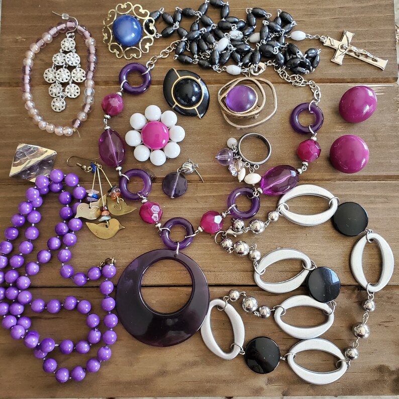Op de afbeelding: Een verzameling sieraden met paarse, witte en zwarte kralen, een zilveren bedel en een zilveren kruis hanger. De sieraden zijn gerangschikt op een houten oppervlak.