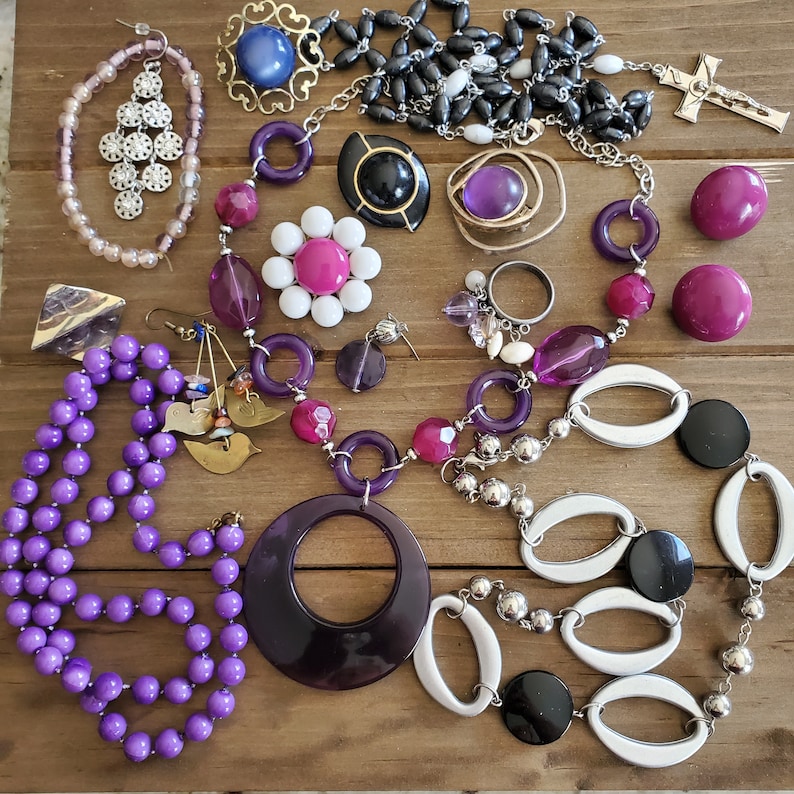 Op de afbeelding: Een verzameling paarse, witte en zwarte sieraden met kralen, hangers en bedels. De sieraden omvatten een ketting met een grote paarse hanger, een armband met witte en zilveren kralen en oorbellen met paarse en witte kralen.