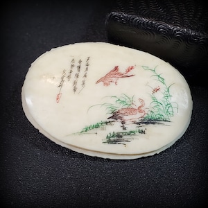 May include: An oval-shaped, white, carved bone pendant with a painted scene of two birds and green foliage. The pendant has Chinese calligraphy written in black ink.