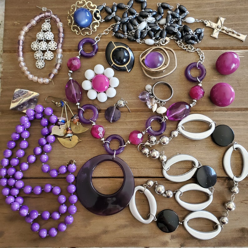 Op de afbeelding: Een verzameling paarse, witte en zwarte sieraden met kralen, hangers en bedels. De sieraden omvatten een ketting met een grote paarse hanger, een armband met witte en zilveren kralen en oorbellen met paarse en witte kralen.