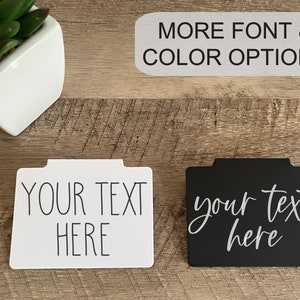 Puede incluir: Dos portaetiquetas rectangulares, uno blanco y otro negro, con el texto "YOUR TEXT HERE" en diferentes fuentes. El portaetiquetas blanco tiene una fuente de bloque, mientras que el portaetiquetas negro tiene una fuente cursiva. También es visible el texto "MORE FONT & COLOR OPTIONS".