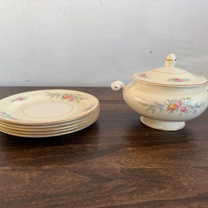 May include: A cream-coloured ceramic serving bowl with a lid and two handles, alongside a stack of matching saucers. Each piece is adorned with a floral pattern in pink, blue, and yellow, with gold trim. The set is displayed on a dark wooden surface.