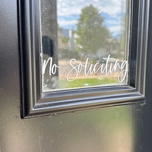 May include: A black door with a glass window displaying the words "No Soliciting" in a cursive white font. The door frame is also black, and the background shows a glimpse of a neighborhood.