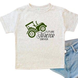 May include: A light heather gray short-sleeve t-shirt featuring a dark green graphic of a tractor and the black text "FUTURE TRACTOR DRIVER" printed on the front. The shirt is paired with blue denim fabric visible at the bottom right corner.