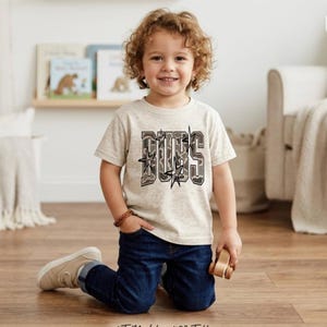 May include: A light beige t-shirt with the word "BUBS" in a camouflage design. The shirt is worn by a young child, paired with blue jeans and tan shoes. The child is smiling and holding a small wooden toy car.
