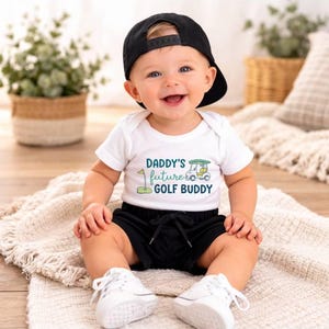 May include: A smiling baby wearing a white onesie with the text "DADDY'S future GOLF BUDDY", black shorts, a black baseball cap, and white sneakers. The baby is sitting on a cream-colored blanket.