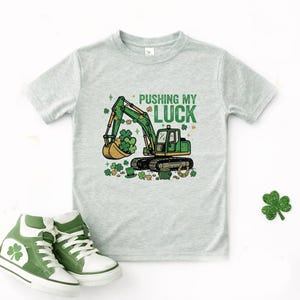 May include: A light gray t-shirt featuring a green excavator scooping up shamrocks, with the text "PUSHING MY LUCK" in green. The shirt is paired with green and white high-top sneakers, perfect for St. Patrick's Day.
