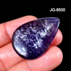 May include: A teardrop-shaped cabochon gemstone with a deep purple hue and internal white, cloud-like inclusions. The surface is polished and smooth, reflecting light across its curved face. The text JG-8500 is visible in the upper right corner.