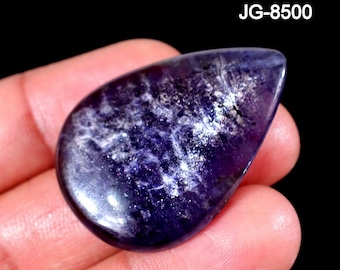 May include: A teardrop-shaped cabochon gemstone with a deep purple hue and internal white, cloud-like inclusions. The surface is polished and smooth, reflecting light across its curved face. The text JG-8500 is visible in the upper right corner.