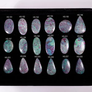 May include: A collection of eighteen polished ruby and zoisite cabochons arranged in three rows on a black display tray. Each gemstone features a unique mottled pattern of magenta, green, and black, labeled with codes from HS-191 to HS-208.