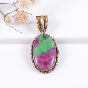 Puede incluir: Colgante ovalado con una piedra de rubí zoisita. La piedra presenta una mezcla de colores verde y magenta, engastada en un marco de metal plateado y dorado. El colgante tiene un diseño de cuerda trenzada alrededor de la piedra y una fianza dorada.