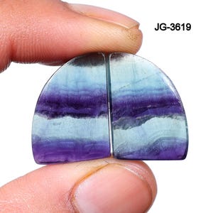 May include: Two polished, semi-circular gemstones, held between fingers, showcasing a blend of purple, blue, and white hues. The stones have a layered appearance, with the text "JG-3619" visible.