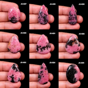 May include: Nine polished rhodochrosite gemstones in various shapes, including oval and teardrop. The stones feature a vibrant pink hue with black and gray veining. Each stone is held in a hand against a black background.