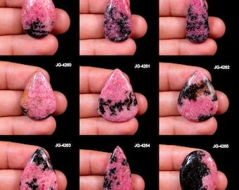 Könnte beinhalten: Neun polierte Rhodochrosit-Edelsteine in verschiedenen Formen, darunter oval und tropfenförmig. Die Steine weisen eine leuchtend rosa Farbe mit schwarzen und grauen Adern auf. Jeder Stein wird in einer Hand vor schwarzem Hintergrund gehalten.