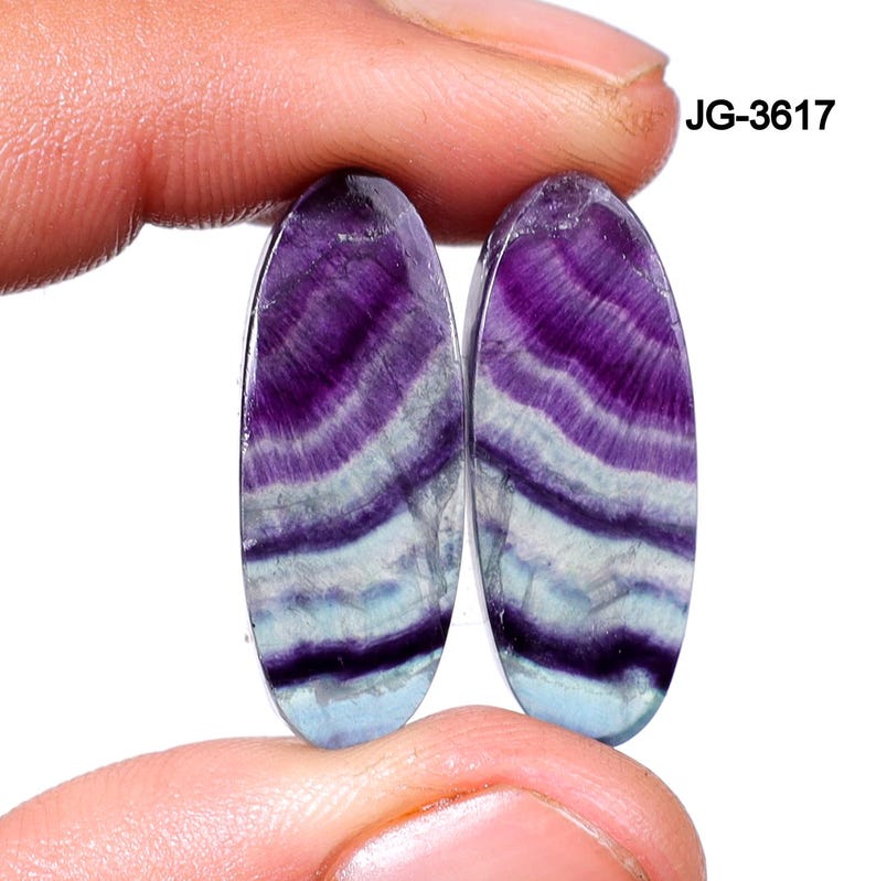 May include: Two oval-shaped gemstones with a layered pattern of purple, white, and blue hues. The gemstones are held between fingers, showcasing their polished surfaces and natural color variations. The text "JG-3617" is visible.