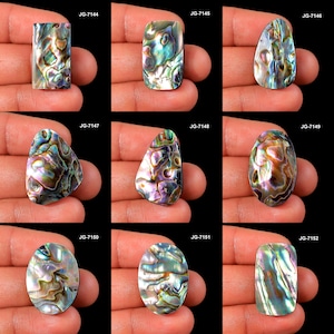 May include: Nine iridescent abalone shell cabochons in diverse shapes, such as rectangular and oval. Each shell showcases a unique pattern of blues, greens, purples, and golds. The cabochons are held in a hand against a black backdrop. Measurements vary.