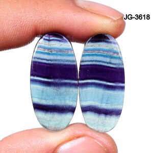 May include: Two oval-shaped gemstones with a layered pattern of purple, light blue, and white. The stones are held between fingers against a white background. The text "JG-3618" is visible in the upper right corner.