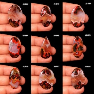 May include: Nine polished gemstone cabochons in various oval and teardrop shapes. The stones feature a mix of colors, including red, brown, white, and orange, with unique patterns and textures. Each stone is held in a hand against a black background.