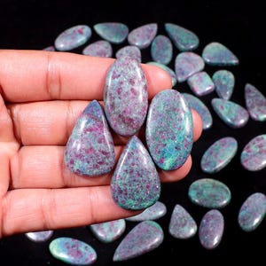 May include: A collection of polished gemstones in various shapes, including oval and teardrop. The stones display a mix of colors, with a dominant teal and magenta pattern. The gemstones are held in a hand, with more scattered in the background.
