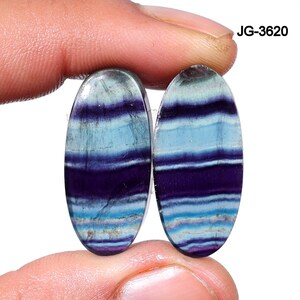 May include: Two oval-shaped gemstones with a layered pattern of blue, purple, and white. The gemstones are held by a person's fingers. The text "JG-3620" is in the upper right corner.